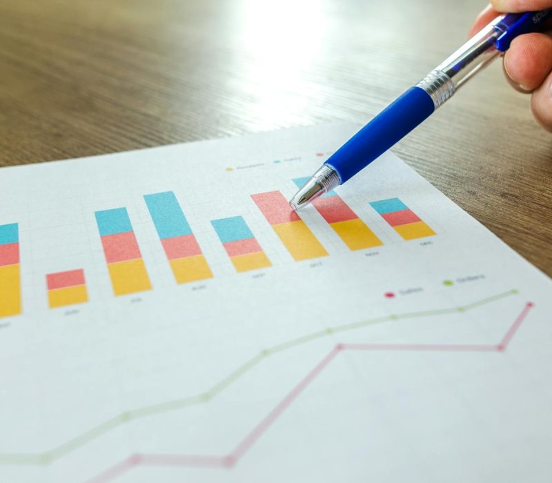 Detailed close-up of a hand pointing at colorful charts with a blue pen on wooden surface.